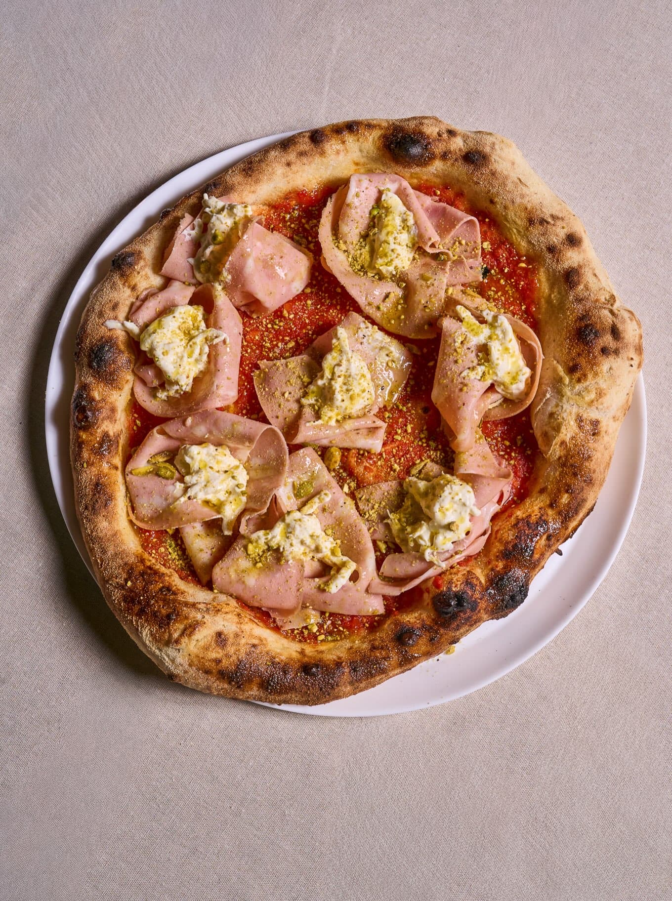 Pizza Mortadella & Pistacchio inteira, com stracciatella e pistache, sobre a mesa de travertino