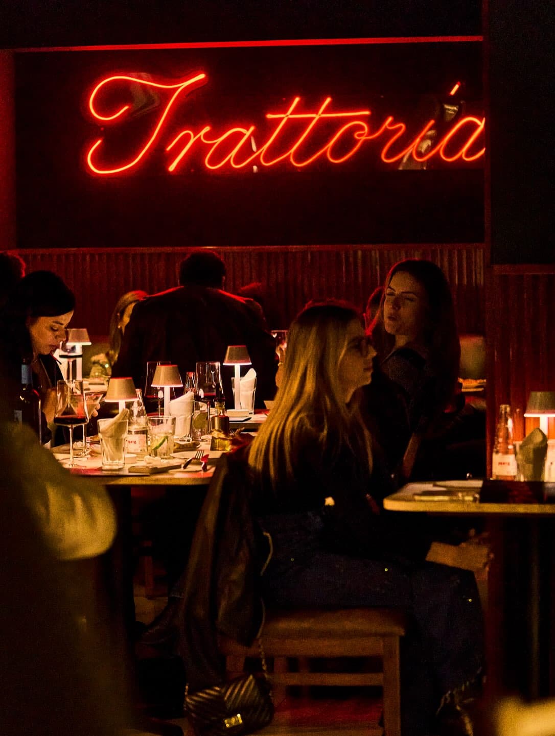 O salão à noite, com o letreiro de neon Trattoria sobre as mesas e as luminárias acesas