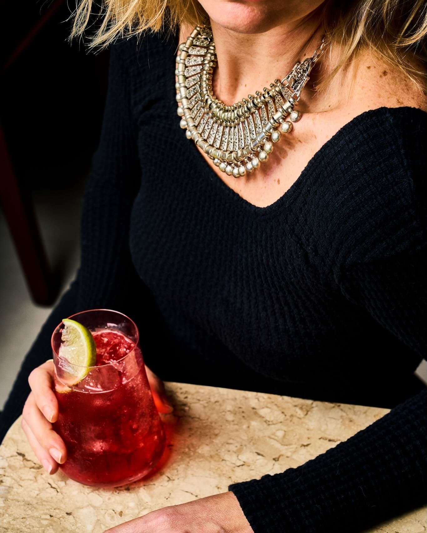 Cliente à mesa de travertino com um drink vermelho na mão, à meia-luz do salão