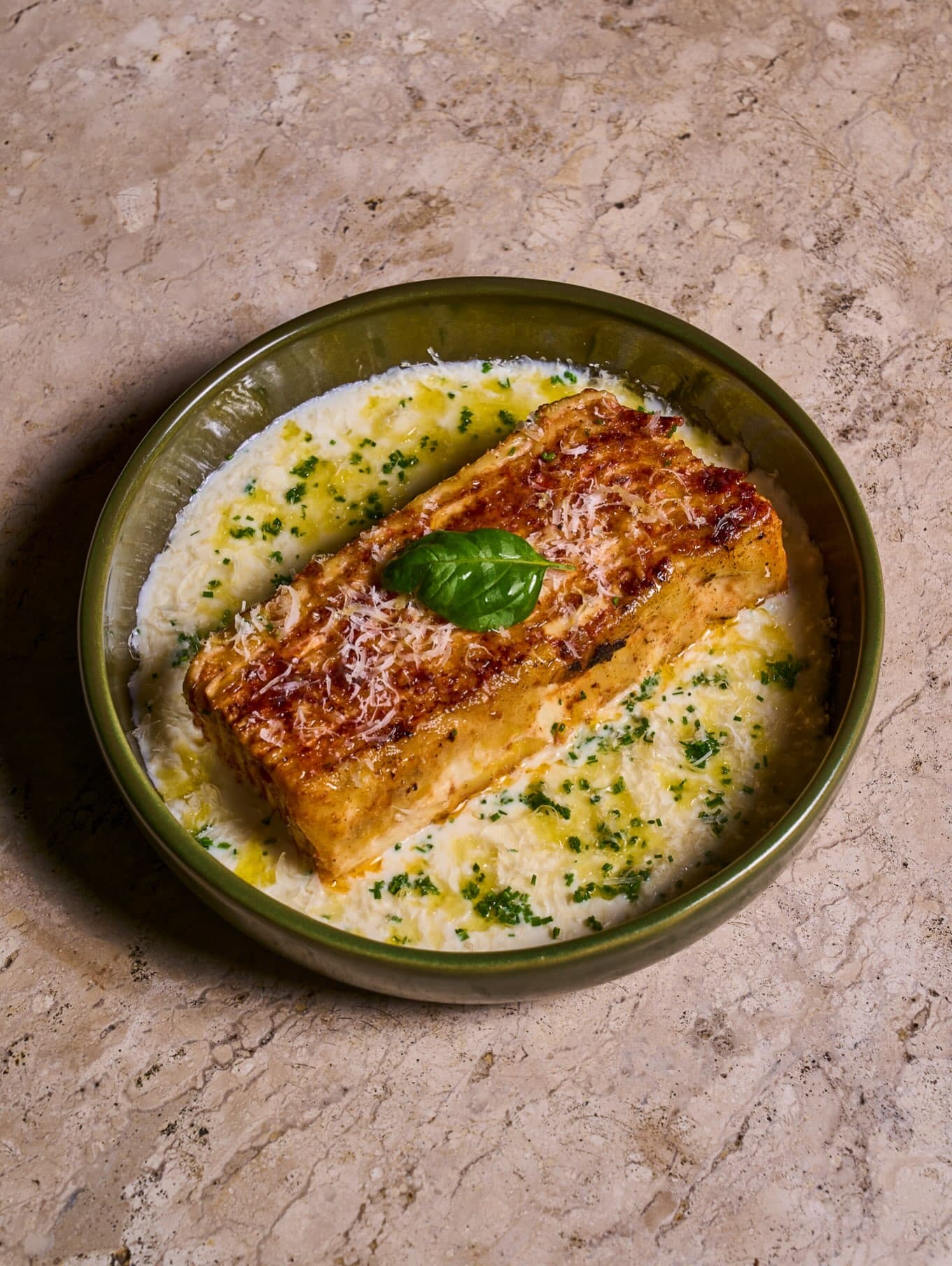 Lasanha Lá Nuova gratinada com folha de manjericão, em prato fundo verde sobre o travertino
